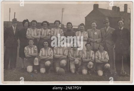 Foto des Zweiten Weltkriegs einer Frauen-Fußballmannschaft. Die Fußballerinnen tragen Shorts und gestreifte Hemden, drei Männer in Anzügen und zwei Frauen in nicht-sportlicher Kleidung stehen zu ihnen. Mehrere Häuser und die Flutlichter eines Fußballplatzes sind im Hintergrund zu sehen Stockfoto