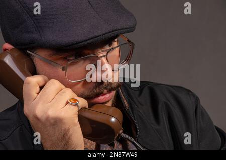 Ein ruhiger Mann mit Brille, braunem Hemd und schwarzer Jacke, der jemanden mit einem alten Telefon anruft Stockfoto