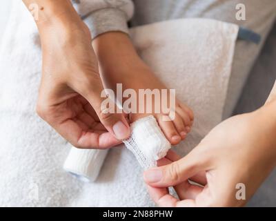 Mutter verbindet den großen Zeh ihres Kindes. Nahaufnahme des Kinderfußes mit Bandfinger. Erste Hilfe bei kleinen häuslichen Verletzungen. Stockfoto