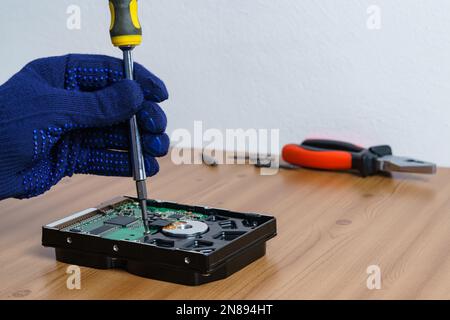 Die Hand eines Mannes in einem Handschuh repariert die Festplatte eines PCs mit einem Schraubendreher. Schließen. Stockfoto