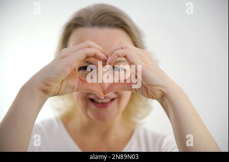 Eine Frau mittleren Alters macht die Hände so, als ob eine Brille vor den Augen das Herz mit den Händen zeigt, mit den Händen durch das Loch schaut Lächeln Liebe Erwartungen Anerkennung Zärtlichkeit Schönheit Hände in Schärfe selektive Schärfe Stockfoto