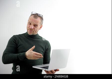 Ein erwachsener Mann bietet beharrlich ein Projekt an, um die Brille auf die Stirn zu heben, um die Hände des Laptops zu halten weißer Hintergrund zeigt die Hand auf technologischem Gerät, das eine zerknitterte Stirn, fururierte Augenbrauen, Bart und Schnurrbart bietet Stockfoto