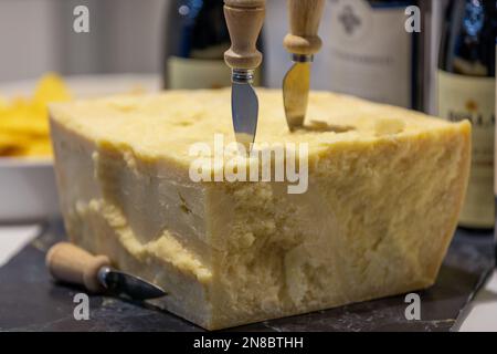 Nahaufnahme des typischen italienischen Parmigiano-Käses mit typischem Messer zum Abnehmen von Stücken, im Hintergrund verschwommene Weinflaschen Stockfoto