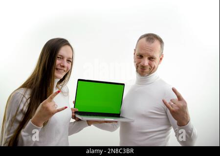 Mann und Mädchen stehen auf weißem Hintergrund in den Händen eines Laptops mit grünem, leerem Bildschirm, zeigen Gesten mit kleinen Fingern und Zeigefinger Ziege magisches Schild schützt vor bösem Ort für zufriedene Werbung Stockfoto