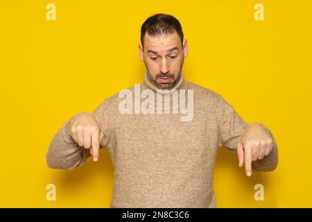 Bärtiger, gutaussehender Latino-Mann mit beigefarbenem Rollkragenpullover, der nach unten zeigt, mit Fingern, die Werbung isoliert auf gelbem Hintergrund zeigen. Stockfoto