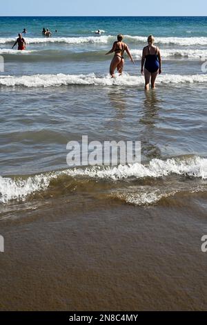 Menschen, die durch leichte Brandung am Sandstrand Playa del Inglés ins Meer gehen, mit anderen im Meer, Maspalomas Stockfoto