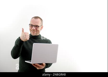 Ein erwachsener Mann mit Brille, Bart und Schnurrbart ist sehr froh, dass er Daumen hoch in den Händen zeigt. Er hat einen grauen Laptop auf weißem Hintergrund 50-60 Jahre alter grüner Pullover viel Glück Erfolg Sieg Stockfoto