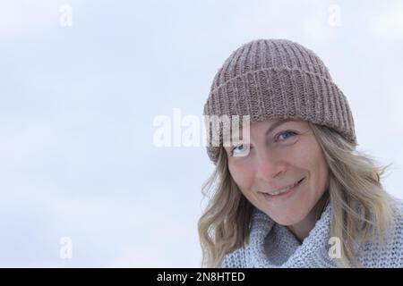 Frauenporträt mit blonden Haaren, Strickmütze und Pullover, lächelnd auf einem leichten Hintergrund Nahaufnahme Stockfoto