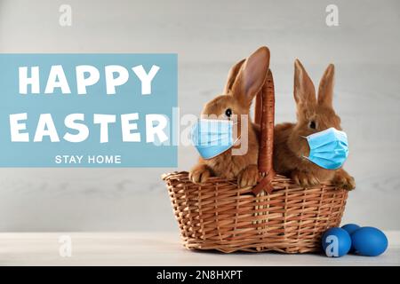 Text Happy Easter Stay Home und niedliche Häschen in Schutzmasken auf dem weißen Tisch. Urlaub während der Covid-19-Pandemie Stockfoto