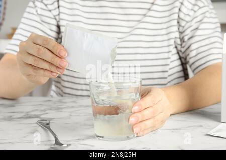 Frau, die Pulver aus dem Arzneimittelbeutel in das Glas mit Wasser am Tisch gießt, Nahaufnahme Stockfoto