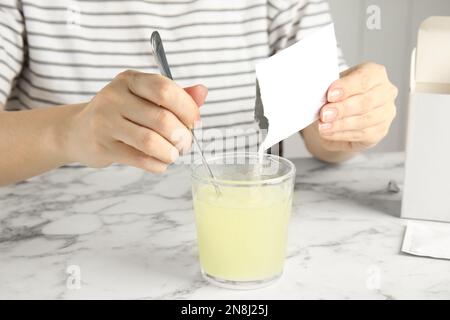 Frau schüttet und rührt Pulver aus dem Arzneimittelbeutel in das Glas mit Wasser am Tisch, Nahaufnahme Stockfoto