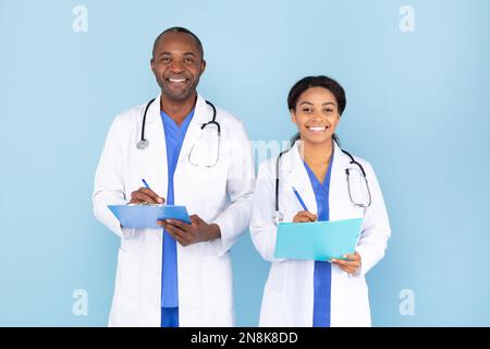 Medizinische Versorgung. Glückliche schwarze, männliche und weibliche Ärzte in Uniform, die Klemmbretter halten und über blauen Hintergrund lächeln Stockfoto