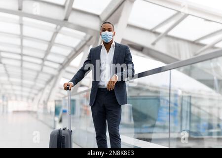 Hübscher Schwarzer Geschäftsmann Trägt Medizinische Schutzmaske, Der Mit Gepäck Am Flughafen Steht Stockfoto