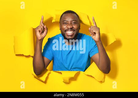 Porträt eines aufgeregten afroamerikanischen Mannes, der mit den Fingern nach oben zeigt und lächelt, der in einem zerrissenen Papierloch auf gelbem Hintergrund steht Stockfoto