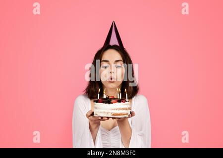 Lächelnde junge europäische Dame mit Hut, Kuchen halten, Kerzen ausblasen, sich etwas wünschen, isoliert auf rosa Hintergrund Stockfoto