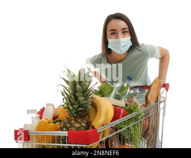 Junge Frau in medizinischer Maske mit Einkaufswagen voller Lebensmittel auf weißem Hintergrund Stockfoto