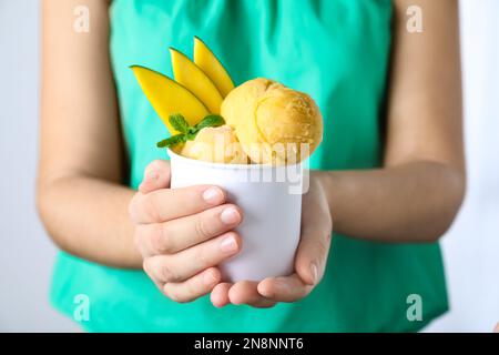 Eine Frau, die eine Schüssel voller köstlicher Eiscreme und Mangoscheiben auf hellgrauem Hintergrund hält, Nahaufnahme Stockfoto