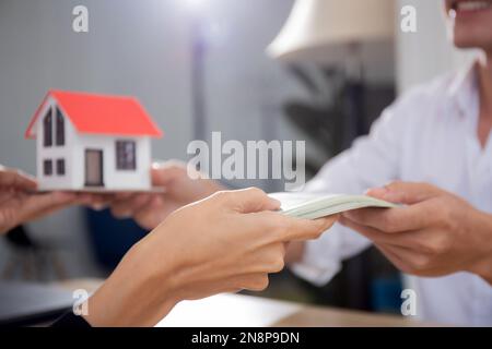 Großhand einer jungen maklerin, die dem Kunden ein Hausmodell überreicht, während der Kunde Geld mit Genehmigung für Immobilien, Investitionen und mehr gibt Stockfoto