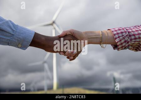 Handschlag zwischen zwei Geschäftsleuten verschiedener ethnischer Herkunft vor Windturbinen. Stockfoto