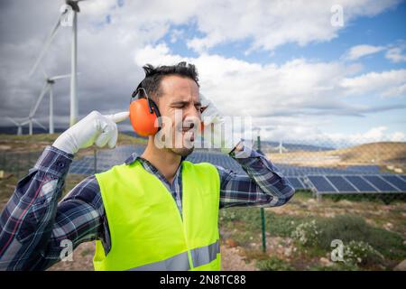 Ein Arbeiter leidet unter Ohrenschmerzen in der Nähe von Windturbinen und trägt daher Kopfhörer mit Geräuschminimierung. Stockfoto