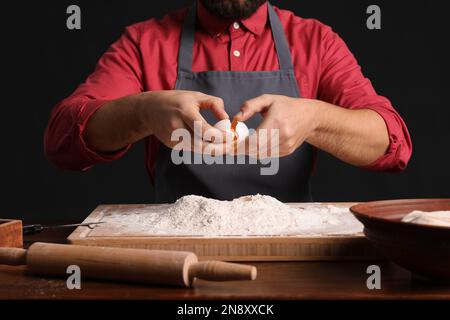 Männlicher Bäcker bricht Ei für Teig am Tisch auf dunklem Hintergrund, Nahaufnahme Stockfoto