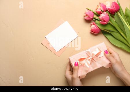 Weibliche Hände halten Geschenk neben Tulpenblumen und leere Karte auf beigefarbenem Hintergrund. Hallo Frühling Stockfoto