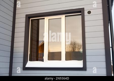 Weißes dreiflügeliges Plastikfenster eines Holzhäuschen in der Wintersaison, das sich im Glas spiegelt Stockfoto