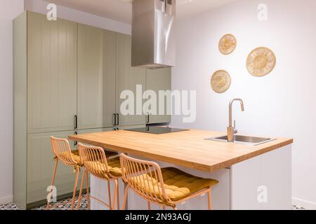 Nahaufnahme des Küchentisches mit trendigen Hochstühlen. Holzplatte und Stühle in einer honigfarbenen Farbpalette. Arbeitsfläche mit eingebautem Spülbecken und Keramik Stockfoto