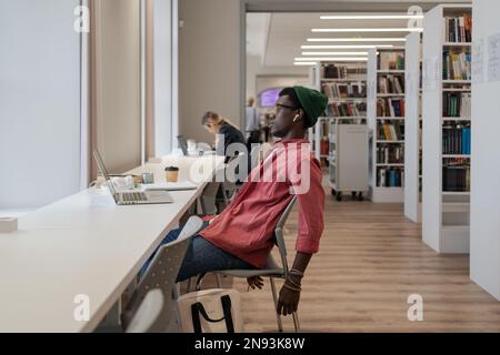 Ein unglücklicher schwarzer Student, der sich vom Lernen überwältigt fühlt, während er online in der Bibliothek lernt Stockfoto