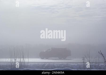 PKW im Nebel auf einer Winterstraße mit großen Schneeverwehungen am Straßenrand. Stockfoto