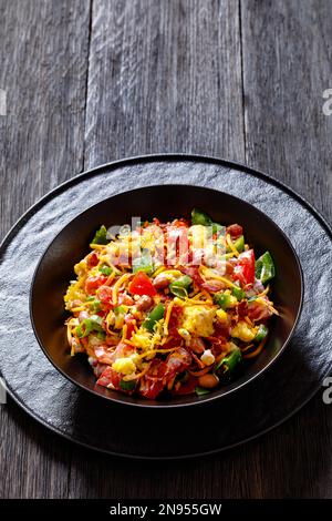 pinto Beans Speck Maisbrotsalat mit Tomaten, Paprika, Mais und Cheddar Käse in schwarzer Schüssel auf dunklem Holztisch, vertikale Sicht von oben Stockfoto