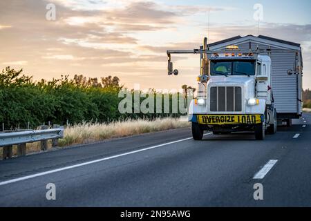 Leistungsstarker, weißer Sattelschlepper für industrielle Schwerlastfahrzeuge mit Übermaß-Lastschild am Stoßfänger für den Transport von übergroßen, gefertigten Klauen Stockfoto