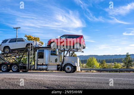 Klassischer Großlaster-Transporter für industrielle Anwendungen Weißer Sattelschlepper, der Autos auf dem modularen hydraulischen Auflieger transportiert, der auf dem schmalen Hang fährt Stockfoto