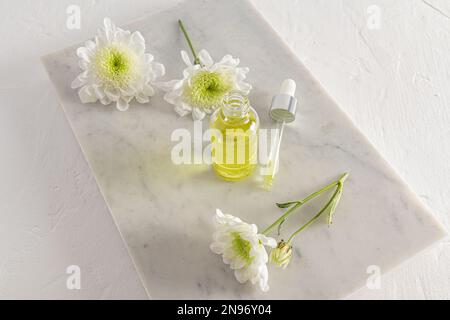 Eine offene Flasche kosmetisches Naturheilmittel und eine gefüllte Pipette auf einem weißen Marmorpodium auf weißem Hintergrund mit Frühlingsblumen. Selbstfürsorge Stockfoto