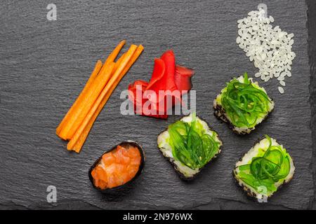 Sushi mit grünen Algen und Lachs. Reis, Ingwer und gehackte Karotten auf dem Tisch. Speicherplatz kopieren. Schwarzer Hintergrund Stockfoto