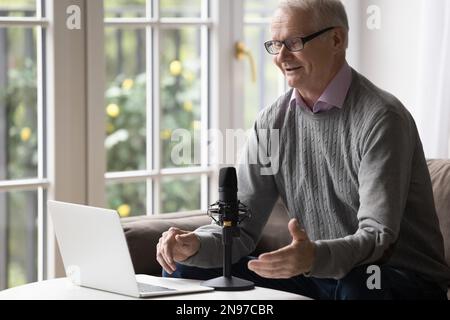 Positiver alter Rentner, der mit Mikrofon und Laptop-Webcam spricht Stockfoto