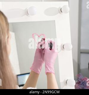 Eine Frau, die medizinische Schutzhandschuhe trägt und ein Herz mit Lippenstift auf einem Spiegel zeichnet. Kosmetikkonzept. Spa und Schönheitssalon Stockfoto