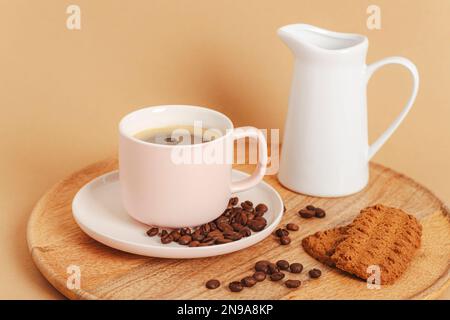 Kaffeetasse, geröstete Kaffeebohnen, Kekse und Kanne mit Milch auf einem Holztablett. Frühstück am Morgen. Nahaufnahme. Stockfoto