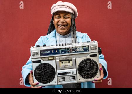 Glückliche, trendige, afrikanische Seniorin, die Spaß beim Musikhören mit klassischer Boombox-Stereoanlage hat Stockfoto