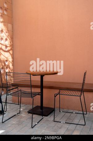 Drei leere schwarze Drahtstühle, eine lange Sitzbank und ein runder Holztisch auf grauen Ziegelsteinböden und ein leerer brauner Terrakotta-Wandhintergrund w Stockfoto