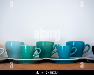 Set aus großen und kleinen blau-grünen Tonnen Kaffeetassen, Keramikbechern mit Untertassen stehen hintereinander auf einem Holzregal isoliert auf weißem Hintergrund mit Kopie sp Stockfoto