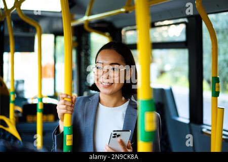 Junge, fröhliche asiatische Frau mit Brille, die während der Busfahrt ein Smartphone in der Hand hält Stockfoto