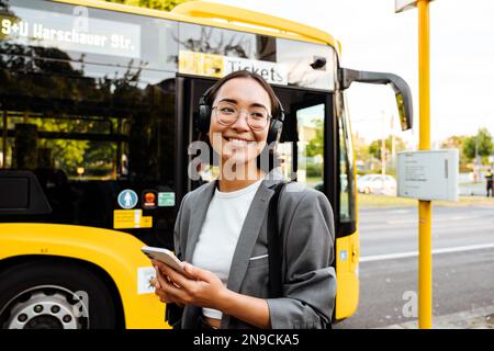 Junge, wunderschöne, lächelnde Asiatin mit Kopfhörern, die ihr Smartphone in der Hand hält, während sie in der Nähe des Busses draußen steht Stockfoto