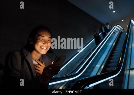 Junge, hübsche asiatische Frau, die ihr Smartphone benutzt und Gesten macht, während sie drinnen auf der Rolltreppe steht Stockfoto
