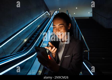 Junge lächelnde asiatische Frau, die Videos und Gesten macht, während sie drinnen auf der Rolltreppe steht Stockfoto