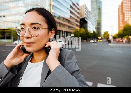 Junge, hübsche asiatische Frau mit Brille, die Kopfhörer abnimmt, während sie auf der Straße steht Stockfoto