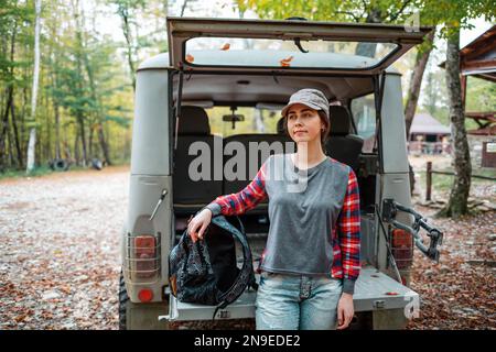 Eine junge weiße Frau posiert mit einem Rucksack gegen den offenen Kofferraum eines suv und schaut weg. Im Hintergrund, Bäume und Wald. Konzept der PKW-Trav Stockfoto