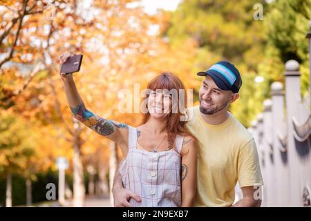 Porträt eines jungen Paares, das auf der Straße posiert und Selfie mit dem Smartphone macht. Glückliche weiße Frau mit Tattoos und einem bärtigen, lächelnden Mann zusammen. Stockfoto