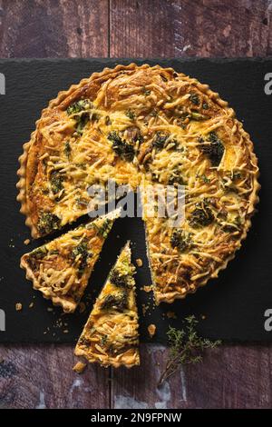 Blick von oben auf eine köstliche Brokkoli-Quiche auf schwarzem Schieferhintergrund auf einem Holztisch, mit zwei ausgeschnittenen Scheiben Stockfoto