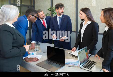 Erfolgreicher Unternehmer, der sich auf die Arbeit mit Diagrammen konzentriert, während er sich mit einem Mitarbeiter aus mehreren ethnischen Gruppen trifft. Reifer Geschäftsmann, der am Konferenztisch über Arbeit diskutiert Stockfoto